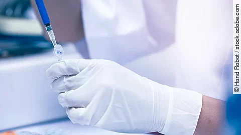 Eine Hand hält ein Eppendorf-Gefäß fest. Mit der anderen Hand wird mit Hilfe einer Spritze und Kanüle Flüssigkeit aus dem Gefäß entnommen. Die Person trägt Handschuhe und sitzt in einem Labor. Vor der Person stehen noch weitere Eppendorf-Gefäße. 