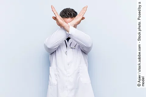 Ein junger Arzt in weißem Kittel versucht sich zu verteidigen indem er seine Arme vor seinem Gesicht verschränkt.