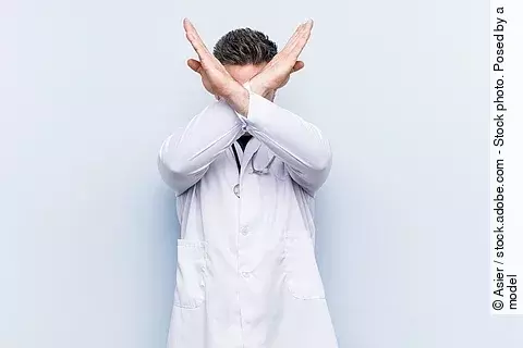 Ein junger Arzt in weißem Kittel versucht sich zu verteidigen indem er seine Arme vor seinem Gesicht verschränkt.