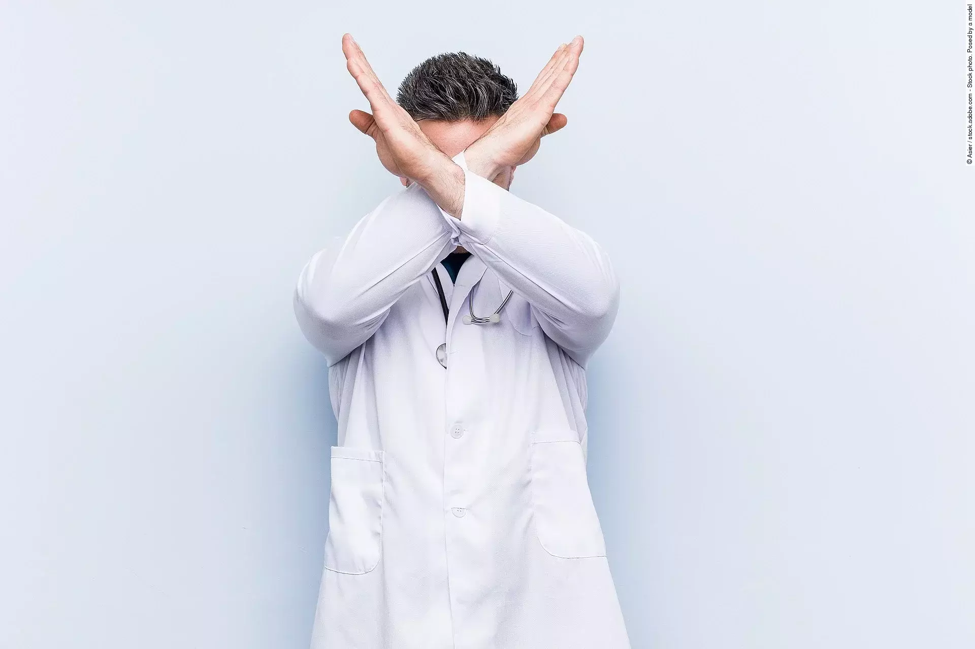 Ein junger Arzt in weißem Kittel versucht sich zu verteidigen indem er seine Arme vor seinem Gesicht verschränkt.