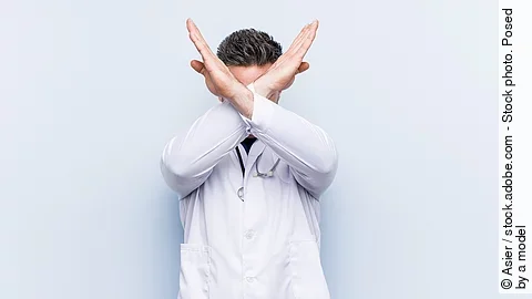 Ein junger Arzt in weißem Kittel versucht sich zu verteidigen indem er seine Arme vor seinem Gesicht verschränkt.