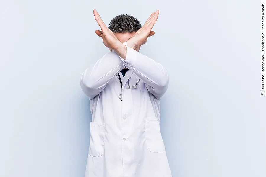 Ein junger Arzt in weißem Kittel versucht sich zu verteidigen indem er seine Arme vor seinem Gesicht verschränkt.
