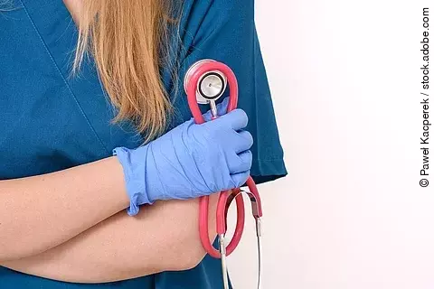 Eine Frau in blauem Kasack steht mit verschränkten Armen vor einem weißen Hintergrund. Sie hat die Arme vor dem Oberkörper verschränkt und trägt blaue Handschuhe. In einer Hand hält sie ein rötliches Stethoskop. Man erkennt nicht das Gesicht, da der Bildausschnitt an den Schultern endet. Die Spitze des Haarzopfs liegt über die Schulter gelegt. .