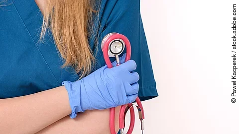 Eine Frau in blauem Kasack steht mit verschränkten Armen vor einem weißen Hintergrund. Sie hat die Arme vor dem Oberkörper verschränkt und trägt blaue Handschuhe. In einer Hand hält sie ein rötliches Stethoskop. Man erkennt nicht das Gesicht, da der Bildausschnitt an den Schultern endet. Die Spitze des Haarzopfs liegt über die Schulter gelegt. .