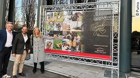 Zwei Männer und eine Frau stehen lächelnd vor einem großen Plakat der Purina-Initiative "Thank you Vets".