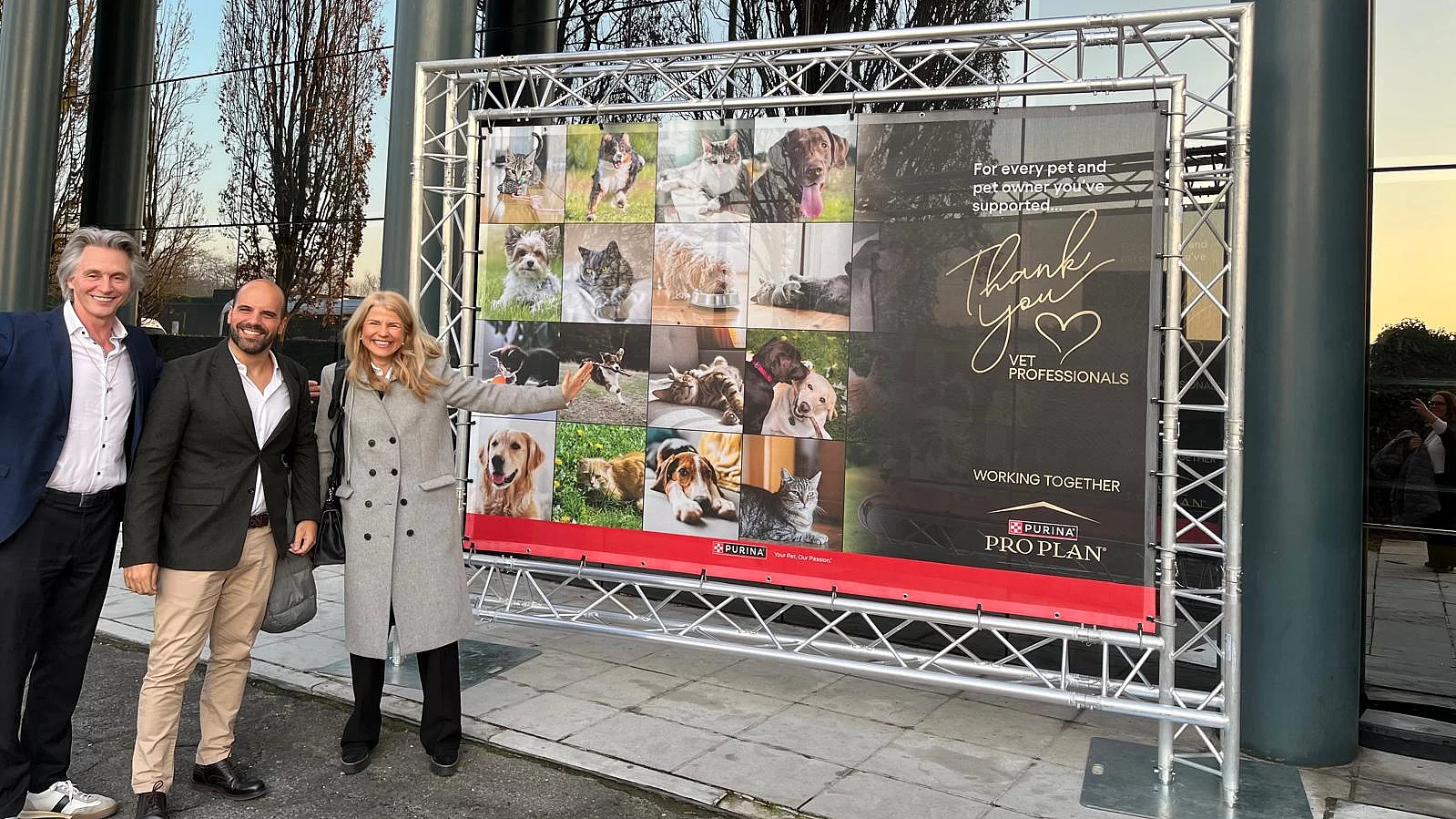 Zwei Männer und eine Frau stehen lächelnd vor einem großen Plakat der Purina-Initiative "Thank you Vets".