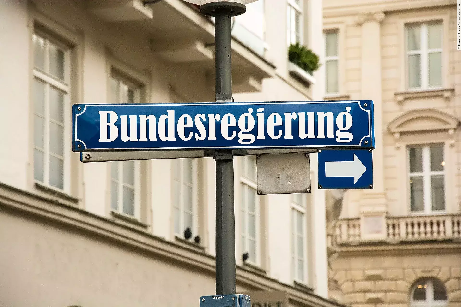Auf einem blauen Straßenschild ist in weißer Schrift "Bundesregierung" geschrieben. Darunter ist ein kleineres Schild mit einem Pfeil zur Seite. Im Hintergrund sind Häuserfassaden.