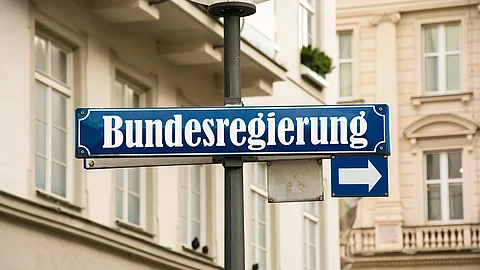 Auf einem blauen Straßenschild ist in weißer Schrift "Bundesregierung" geschrieben. Darunter ist ein kleineres Schild mit einem Pfeil zur Seite. Im Hintergrund sind Häuserfassaden.