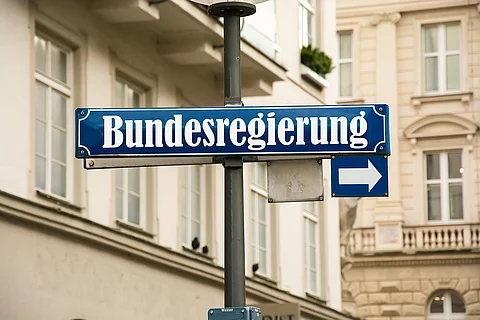 Auf einem blauen Straßenschild ist in weißer Schrift "Bundesregierung" geschrieben. Darunter ist ein kleineres Schild mit einem Pfeil zur Seite. Im Hintergrund sind Häuserfassaden.