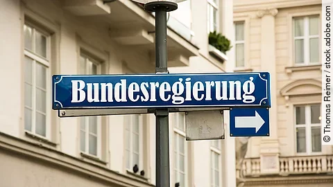 Auf einem blauen Straßenschild ist in weißer Schrift "Bundesregierung" geschrieben. Darunter ist ein kleineres Schild mit einem Pfeil zur Seite. Im Hintergrund sind Häuserfassaden.
