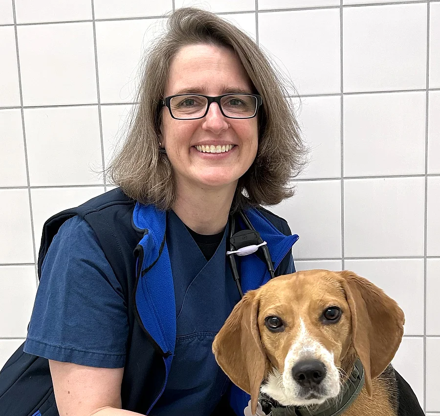 Claudia Richter steht in einem Behandlungszimmer und lacht in die Kamera. Neben ihr sitzt ein Beagle. 