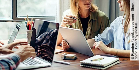 Eine Gruppe junger Menschen arbeitet zusammen an einem Schreibtisch. Die Personen haben Laptops und Schreibutensilien vor sich liegen. Der Schreibtisch steht an einem Fenster, durch das Licht einfällt. Zwei Frauen reden miteinander und ein Mann tippt auf der Tastatur seines Laptops.