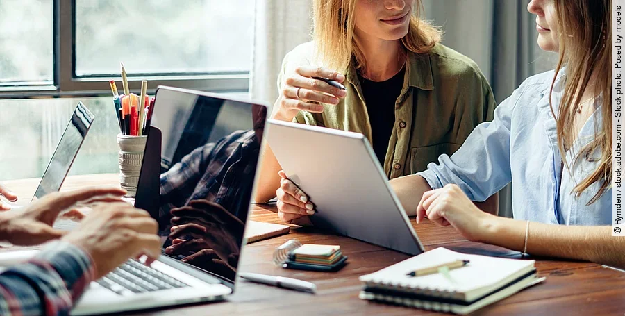 Eine Gruppe junger Menschen arbeitet zusammen an einem Schreibtisch. Die Personen haben Laptops und Schreibutensilien vor sich liegen. Der Schreibtisch steht an einem Fenster, durch das Licht einfällt. Zwei Frauen reden miteinander und ein Mann tippt auf der Tastatur seines Laptops.