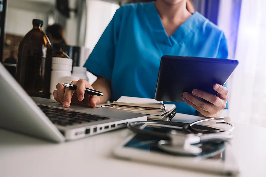 Eine Person sitzt an einem Schreibtisch mit Laptop und Tablet. Die Person trägt einen blauen Kasak. 
