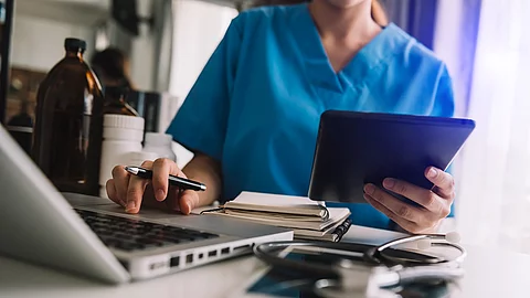 Eine Person sitzt an einem Schreibtisch mit Laptop und Tablet. Die Person trägt einen blauen Kasak. 