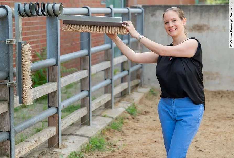 Borbala Foris steht lächelnd in einem Auslauf für Rinder und hält eine Bürste zum Kratzen für die Tiere fest. Dabei sieht sie glücklich aus. 