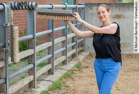 Borbala Foris steht lächelnd in einem Auslauf für Rinder und hält eine Bürste zum Kratzen für die Tiere fest. Dabei sieht sie glücklich aus. 