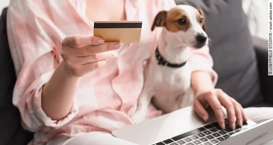 partial view of young woman holding credit card near dog and laptop while online shopping at home, banner Eine Frau sitzt auf einem Sofa. Auf ihrem Schoß sitzt ein kleiner Jack Russell Terrier mit weiß-braunem und glattem Fell. Außerdem hält die Person in der rechten Hand eine Bankkarte. Auf ihren Knien ist ein Laptop abgestellt, wessen sie mit der linken Hand bedient.
