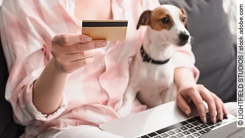 Eine Frau sitzt auf einem Sofa. Auf ihrem Schoß sitzt ein kleiner Jack Russell Terrier mit weiß-braunem und glattem Fell. Außerdem hält die Person in der rechten Hand eine Bankkarte. Auf ihren Knien ist ein Laptop abgestellt, wessen sie mit der linken Hand bedient.