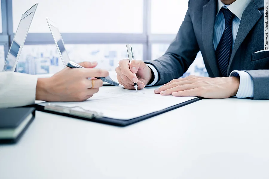 Zwei Personen sitzen sich gegenüber an einem Tisch. Zwischen ihnen liegen Dokumente..