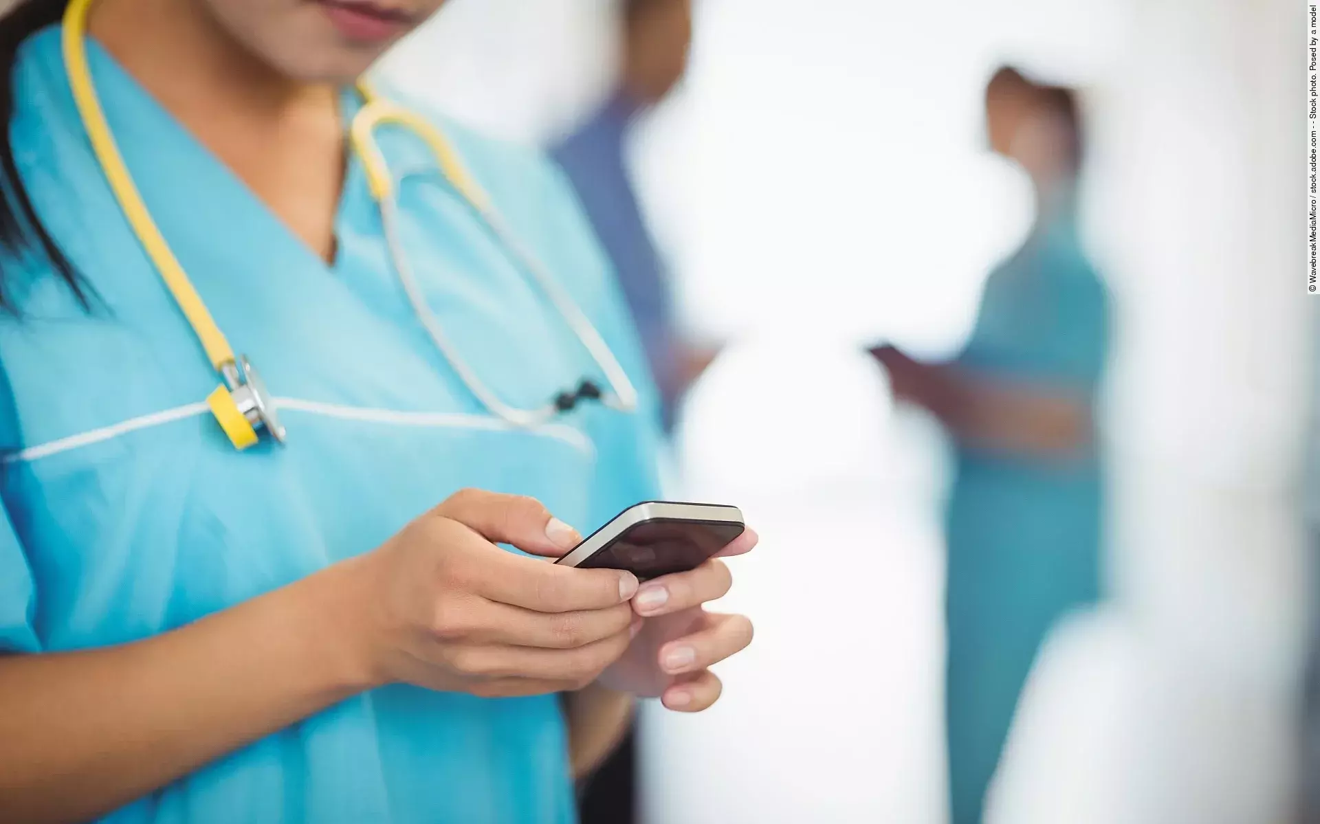 Eine TFA trägt einen blauen Kassak und ein gelbes Stethoskop während sie auf ihr Handy schaut. Im Hintergrund stehen 2 ihrer Kolleg*innen.