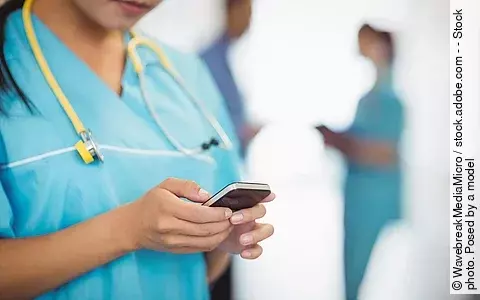 Eine TFA trägt einen blauen Kassak und ein gelbes Stethoskop während sie auf ihr Handy schaut. Im Hintergrund stehen 2 ihrer Kolleg*innen.