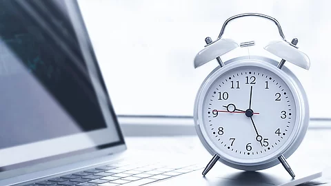 Auf einem Schreibtisch steht ein Laptop. Vor dem Lapto steht ein weißer Wecker. Die Uhrzeit auf dem Wecker ist 12:26 Uhr. Der Laptop ist silber.