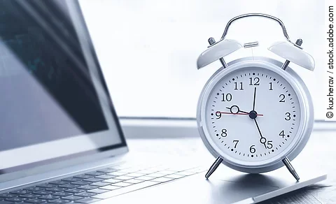 Auf einem Schreibtisch steht ein Laptop. Vor dem Lapto steht ein weißer Wecker. Die Uhrzeit auf dem Wecker ist 12:26 Uhr. Der Laptop ist silber.