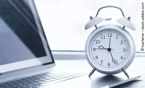 Auf einem Schreibtisch steht ein Laptop. Vor dem Lapto steht ein weißer Wecker. Die Uhrzeit auf dem Wecker ist 12:26 Uhr. Der Laptop ist silber.