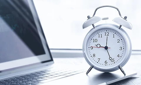 Auf einem Schreibtisch steht ein Laptop. Vor dem Lapto steht ein weißer Wecker. Die Uhrzeit auf dem Wecker ist 12:26 Uhr. Der Laptop ist silber.