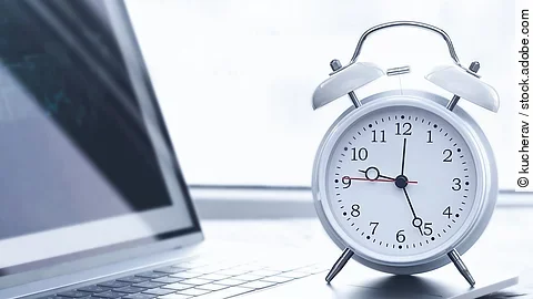 Auf einem Schreibtisch steht ein Laptop. Vor dem Lapto steht ein weißer Wecker. Die Uhrzeit auf dem Wecker ist 12:26 Uhr. Der Laptop ist silber.