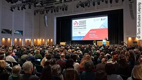 Viele Menschen sitzen in einem abgedunkelten Saal eines Messegeländes und hören einem Mann zu, der vorne auf einer Bühne am Rednerpult steht.