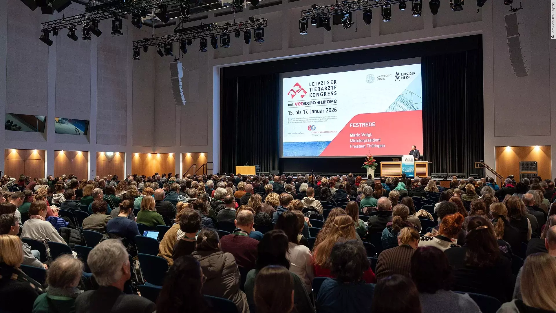 Viele Menschen sitzen in einem abgedunkelten Saal eines Messegeländes und hören einem Mann zu, der vorne auf einer Bühne am Rednerpult steht.