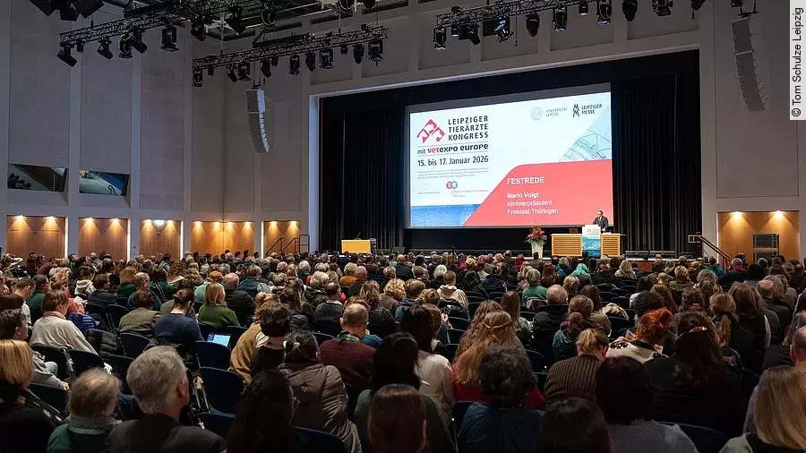 Viele Menschen sitzen in einem abgedunkelten Saal eines Messegeländes und hören einem Mann zu, der vorne auf einer Bühne am Rednerpult steht.