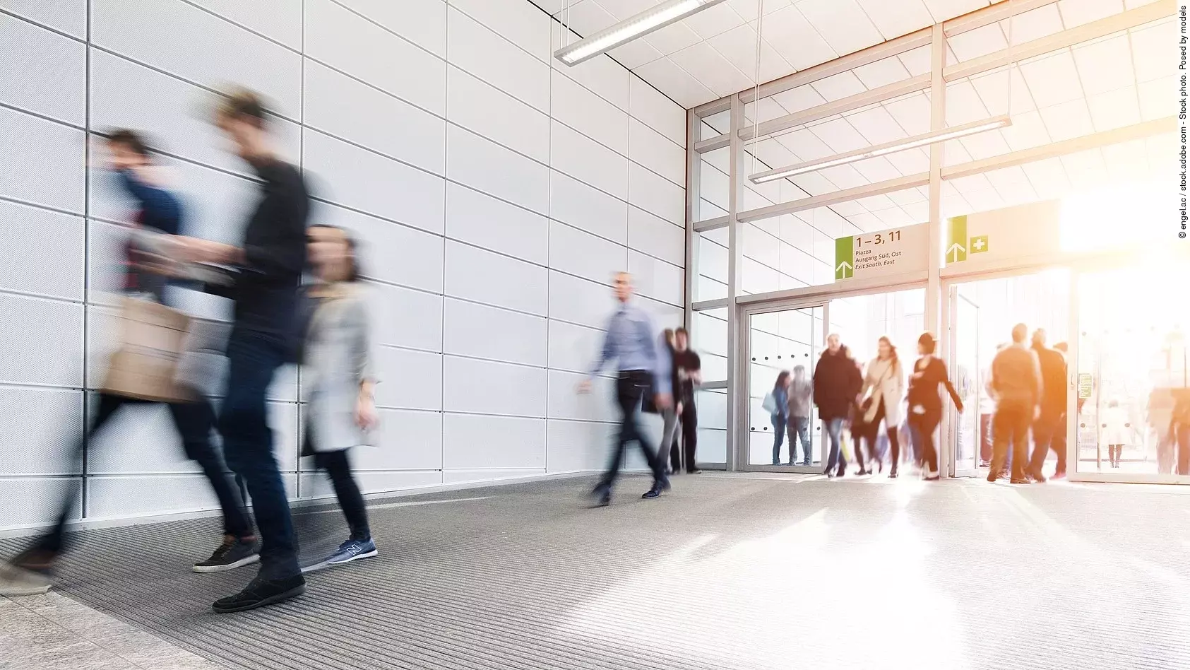 Menschen laufen im Korridor einer Messehalle. Das Bild ist verschwommen. Die Personen tragen formelle Kleidung. 