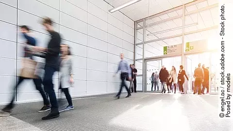 Menschen laufen im Korridor einer Messehalle. Das Bild ist verschwommen. Die Personen tragen formelle Kleidung. 