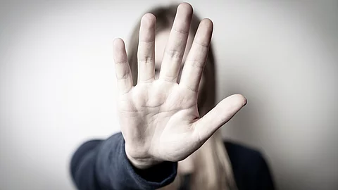 Eine Frau hält ihre aiusgestrekcte Hand vor sich und verdeckt damti ihr Gesicht. Sie trägt ein schwarzes Langarmshirt. Das Bild ist in graulichen Farben gehalten.
