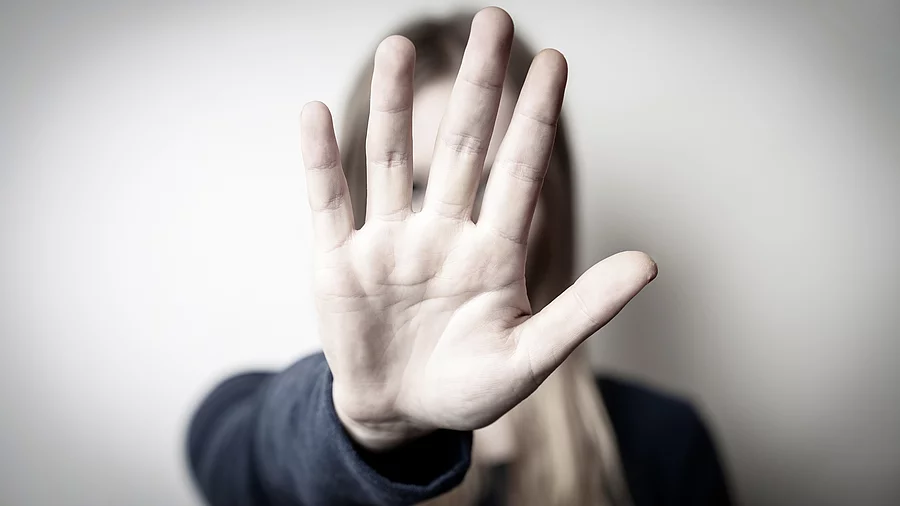 Eine Frau hält ihre aiusgestrekcte Hand vor sich und verdeckt damti ihr Gesicht. Sie trägt ein schwarzes Langarmshirt. Das Bild ist in graulichen Farben gehalten. 