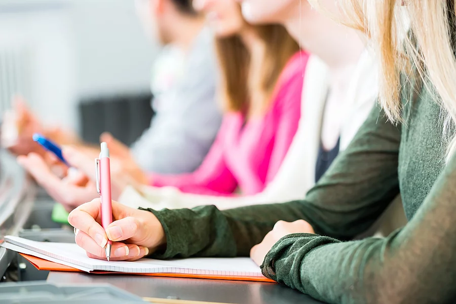 Students in college learning Vier Studierende sitzen in einer Reihe an einem Tisch. Man erkennt nur die Oberkörper der Studierenden. Sie halten Stifte in der Hand und haben Notizen vor sich ausgebreitet.