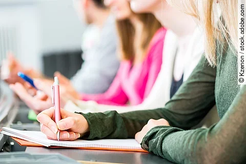 Vier Studierende sitzen in einer Reihe an einem Tisch. Man erkennt nur die Oberkörper der Studierenden. Sie halten Stifte in der Hand und haben Notizen vor sich ausgebreitet.