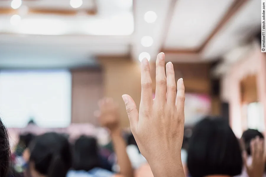 Audience or students raising hands up at conference to answer qu Die gehobene Hand einer Person ist im Fokus des Bildes. Im Hintergrund sieht man verschwommen einige Menschen auf Stühlen sitzend in einem Konferenzraum. Außerdem ist eine Präsentation auf einer großen Wand projiziert, in diese Richtung schauen die Teilnehmer*innen.
