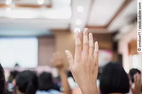 Die gehobene Hand einer Person ist im Fokus des Bildes. Im Hintergrund sieht man verschwommen einige Menschen auf Stühlen sitzend in einem Konferenzraum. Außerdem ist eine Präsentation auf einer großen Wand projiziert, in diese Richtung schauen die Teilnehmer*innen.
