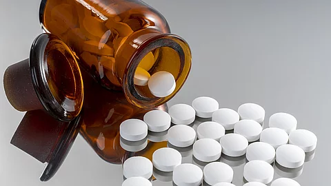 Weiße Tabletten vor einer liegenden Tablettenflasche aus Glas. Auf einer silbernen Unterlage liegen weiße Tabletten. Außerdem liegt eine umgekippte Tablettenflasche hinter den Tabletten. In der Flasche befinden sich auch noch weiße Tabletten. Die Flasche ist aus dunklem Glas.
