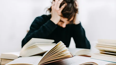 Eine Studentin sitzt verzweifelt am Schriebtisch. Vor ihr liegen einige aufgeschlagene Bücher.