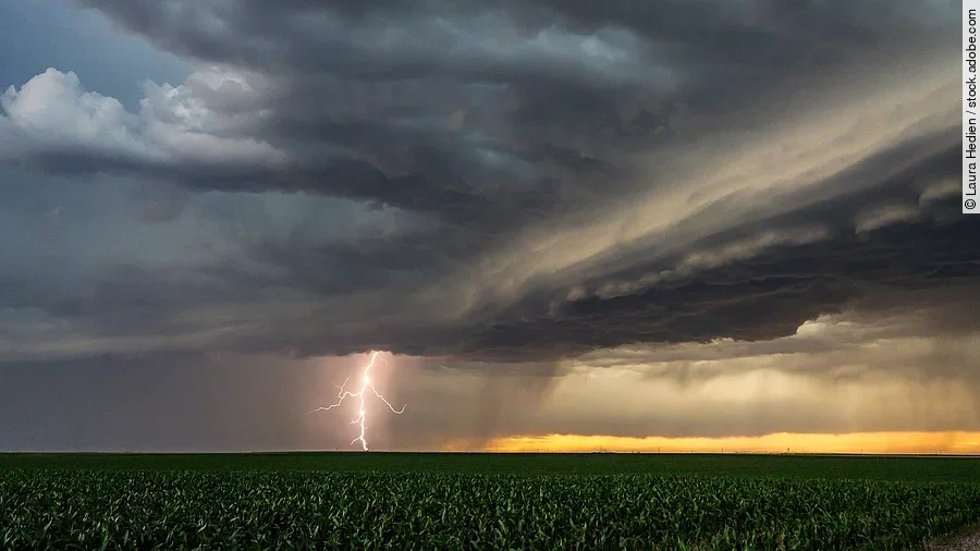 Blitze, Donner und Unwetter in den Great Plains. Ein Gewitter ist über einem Feld in den Great Plains. Ein Blitz zieht von den Wolken Richtung Erde. Die Wolken sind dunkel. In weiter Ferne ist der Himmel wird heller in einem gelblichen Ton.