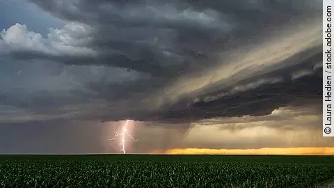 Ein Gewitter ist über einem Feld in den Great Plains. Ein Blitz zieht von den Wolken Richtung Erde. Die Wolken sind dunkel. In weiter Ferne ist der Himmel wird heller in einem gelblichen Ton. 