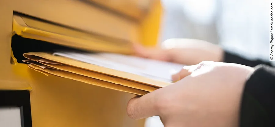 Eine Person steckt ein paar große Briefe in einen Breifkasten. Der Briefkasten ist gelb. Die Person trägt ein schwarze langärmliges Oberteil.