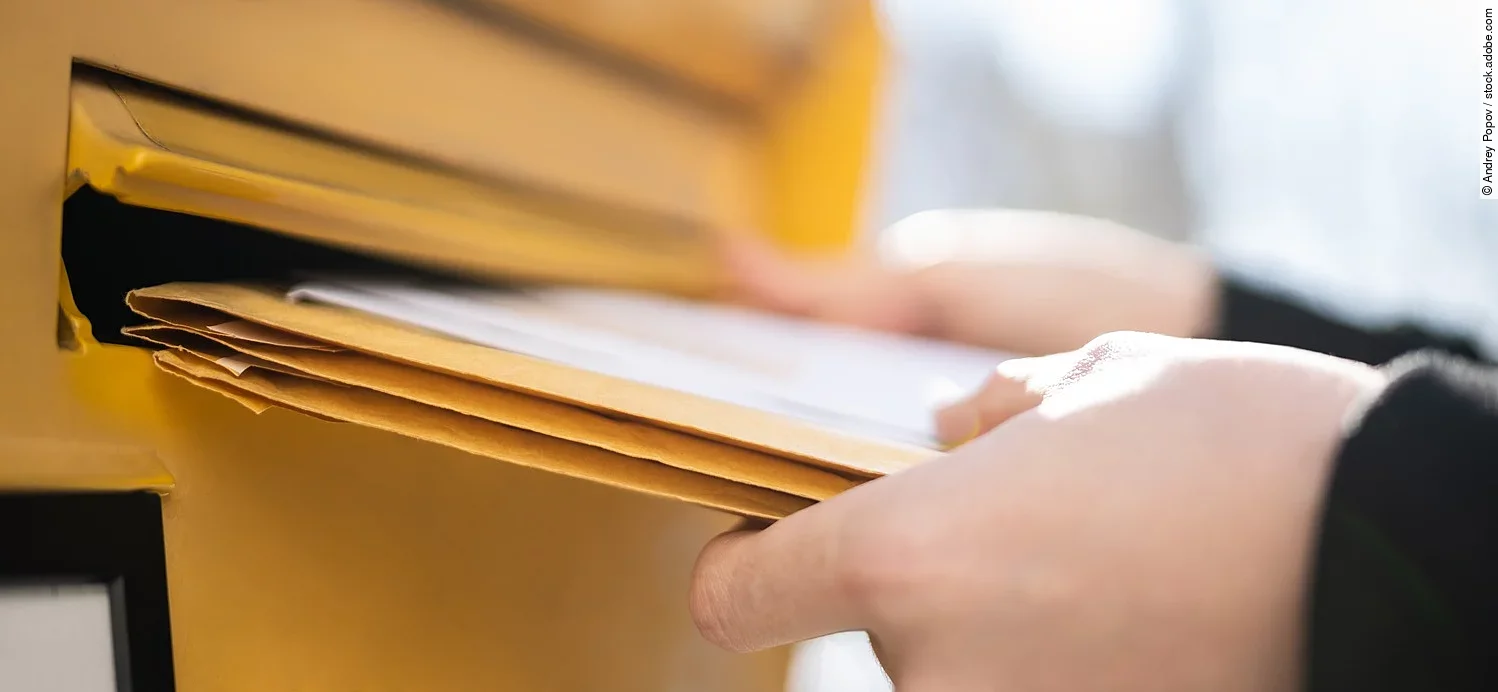 Eine Person steckt ein paar große Briefe in einen Breifkasten. Der Briefkasten ist gelb. Die Person trägt ein schwarze langärmliges Oberteil.