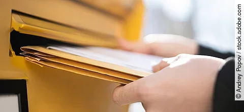 Eine Person steckt ein paar große Briefe in einen Breifkasten. Der Briefkasten ist gelb. Die Person trägt ein schwarze langärmliges Oberteil.