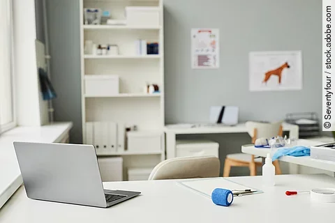 Arbeitsplatz in einer Tierarztpraxis. Im Vordergrund des Bildes steht ein weißer Schreibtisch. Auf dem Tisch ist ein geöffneter Laptop abgestellt und weitere Utensilien - wie eine Bandage und ein Klemmbrett - liegen auf dem Tisch. Im Hintergrund steht eine Bücherregal und medizinische Plakate hängen an der Wand.